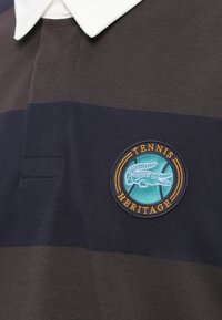Tumed sinise ja halli triibuga polo särk, millel on valge krae ja ümmargune logo koos sõnadega "Tennis Heritage" ning krokodillikujutisega.