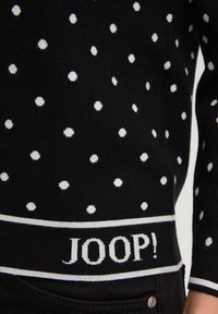 Μαύρο πουλόβερ με λευκές πουά, ριμπ ύφανση και αντίθετη μέση με την επιγραφή "JOOP!" σε λευκά γράμματα.