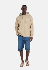 Beige Baumwoll-Hoodie mit Kordeln und Kängurutasche, kombiniert mit dunklen Denim-Shorts. Die Schuhe weisen hellbraune und beige Akzente auf.
