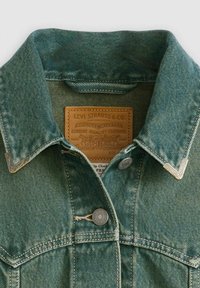 Denim jacket in faded teal with a pointed collar, silver hardware buttons, and a leather label featuring brand details.