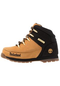 Bota de senderismo en tonos marrón y negro, hecha de materiales de cuero y textil, con ojales dorados, un cuello texturizado y el logo de Timberland en el lateral.