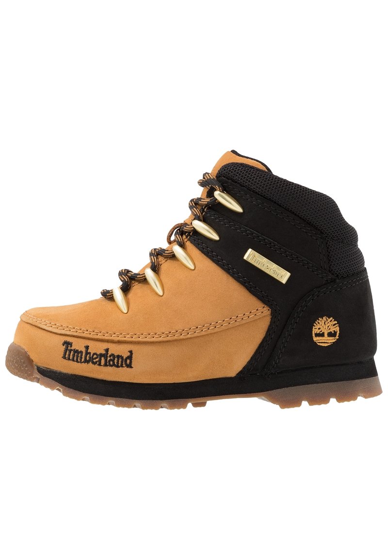 Bota de senderismo en tonos marrón y negro, hecha de materiales de cuero y textil, con ojales dorados, un cuello texturizado y el logo de Timberland en el lateral.
