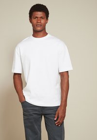 Man met kort krullend haar die een effen wit T-shirt met korte mouwen en donkergrijze spijkerbroek draagt, staat met één hand in zijn zak.