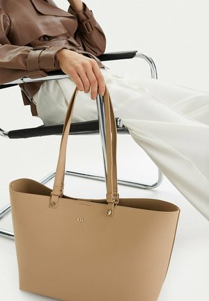 Person sitzt auf einem modernen Stuhl, trägt eine braune Lederjacke und weiße Hosen und hält eine große beige Tragetasche mit goldenen Details.