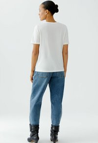 Weißes T-Shirt mit kurzen Ärmeln, kombiniert mit blauen, locker sitzenden Jeans und schwarzen knielangen Stiefeln, von hinten betrachtet. Glattes Material mit klaren Linien.