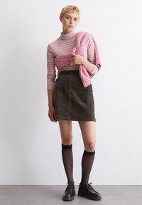 Enganzter, gestreifter Rollkragenpullover in Pink und Weiß; olivgrüner Cordrock; schwarze Kniestrümpfe; schwarze Schnürschuhe; rosa Pullover über die Schultern drapiert.