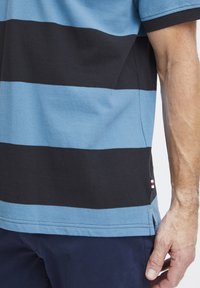 Pruhovaný polo tričko s střídavými vodorovnými pruhy světle modré a černé barvy, ribbovaný límeček a malou štítkovou nášivku na lemu.