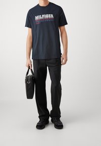 Camiseta de algodón azul marino con el logo "HILFIGER" en rojo y blanco, combinada con pantalones negros y un bolso de cuero negro. Zapatillas deportivas negras gruesas.
