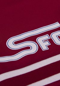 Bordeauxkleurige stof met witte en blauwe gestreepte accenten en geborduurde letters "SFC" in een golvend ontwerp. Gestructureerd, stevig materiaal.