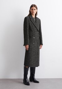 Cappotto lungo doppiopetto in tweed nero e grigio con finitura testurizzata, dotato di due tasche frontali e bottoni scuri, abbinato a stivali neri.