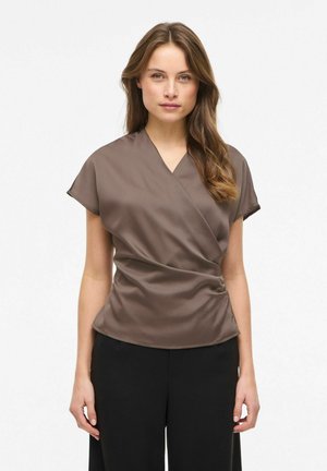 Femme aux longs cheveux bruns portant un chemisier taupe à manches courtes de style cache-cœur et un pantalon noir, debout devant un fond blanc.