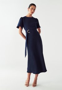 Námornícky modré šaty s krátkymi puff rukávmi, stiahnutý pás s pásikom zo zlatého krúžku a splývavý A-číslovaný strih. Hladký povrch.