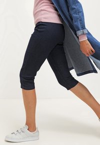 Persona vistiendo pantalones oscuros hasta la rodilla, zapatillas blancas, un suéter rosa y una larga chaqueta de mezclilla azul caminando.