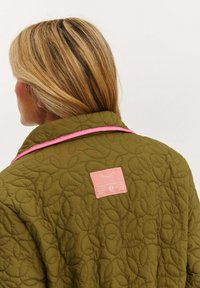 Olivovo zelená prešívaná bunda s ružovým golierom a obdĺžnikovou ružovou štítkom s textom. Textúra obsahuje vzory pripomínajúce listy.