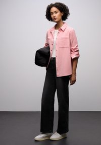 Różowa koszula zapinana na guziki z dwoma przednimi kieszeniami, biała koszulka pod spodem, czarne szerokie spodnie dżinsowe i białe trampki, trzymająca czarną kurtkę.