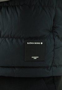 Tamno plava puffer jakna s teksturiranom površinom. Ima crnu oznaku s bijelim tekstom "BJÖRN BORG STOCKHOLM 1956" ušivenom na njoj.