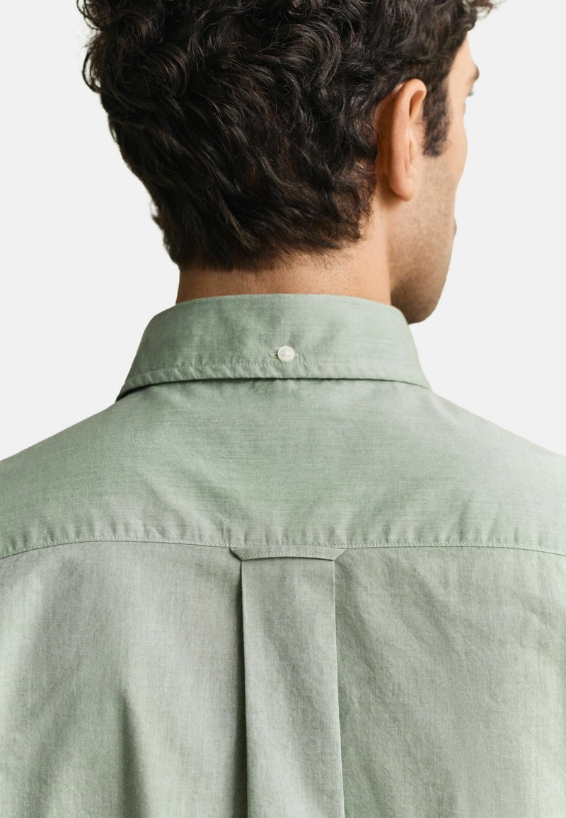 Hombre con cabello corto y rizado que lleva una camisa verde claro con cuello, visto desde atrás mostrando la espalda con canesú y detalles de pliegue central.