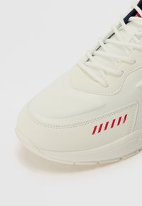 White athletic shoe featuring a textured upper, mesh detailing, and subtle red striped accents on the side. Flat laces and cushioned sole.
