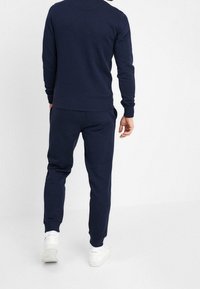 Man som bär en marinblå sweatshirt och matchande joggingbyxor med vita sneakers, vänd bort från en enfärgad vit bakgrund.