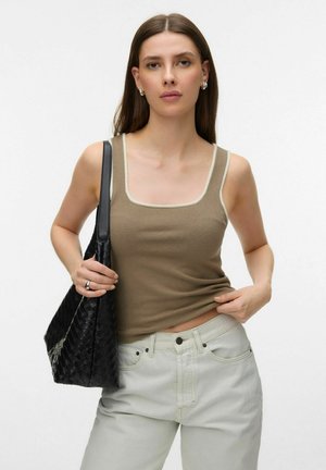 Femme portant un débardeur marron et un pantalon clair, tenant un sac à main noir tissé sur son épaule, sur un fond uni.