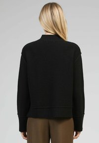 Femme blonde tournant le dos, portant un pull en tricot noir à côtes et un pantalon marron, sur un fond gris uni.