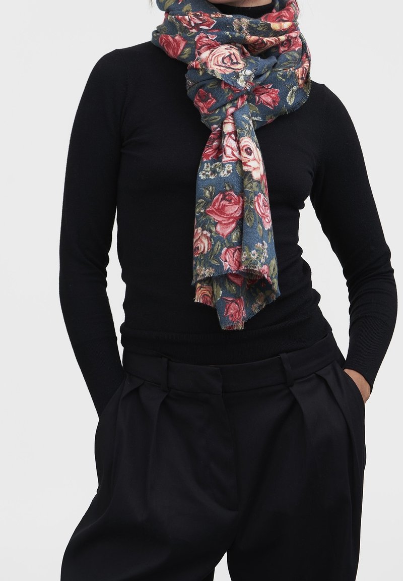 Personne portant un haut noir à manches longues et un pantalon avec une écharpe bleue à fleurs enroulée et nouée autour du cou, mains dans les poches, sur fond clair.