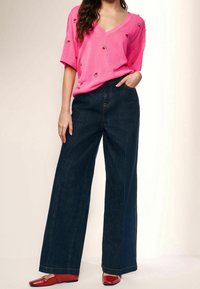 Suéter rosa de cuello en V con un pequeño bordado de sandía, combinado con pantalones de mezclilla oscuros de talle alto y pierna ancha, y unos zapatos planos rojos brillantes.