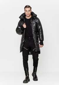 Mężczyzna idący do przodu w błyszczącej czarnej kurtce puchowej z kapturem, czarnych spodniach z zamkami błyskawicznymi i czarnych butach na tle białego tła.