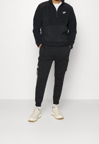 Schwarzer Nike-Polyester-Pullover mit Reißverschluss und Kängurutasche, kombiniert mit schwarzen Jogginghosen mit Cargo-Taschen und weißen Seitenstreifen. Beigefarbene Sneakers sind ebenfalls enthalten.