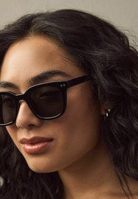 Lunettes de soleil noires avec des montures épaisses, des verres foncés et de petits accents dorés. Cheveux bouclés et foncés partiellement visibles, teint clair.