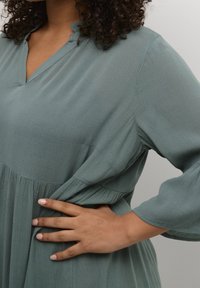 Green, lightweight dress with a V-neckline and flared sleeves. The fabric features a soft texture and pleated detail at the waist.