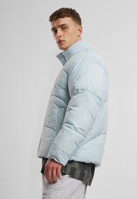 Ljusblå puffjacka med hög krage, quiltad struktur och sidofickor, bärs över en mörk t-shirt med grafiskt mönster och grå sweatpants.