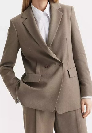 Kobieta w beżowej dwurzędowej marynarce z kieszeniami z klapami, na białej koszuli z kołnierzykiem, w zestawie z beżowymi spodniami.