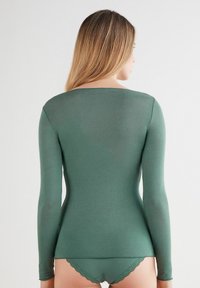 Top de manga larga en verde oscuro, hecho de un material liso y ligero. Presenta un cuello redondo y una silueta ajustada, combinado con bragas a juego con encaje en los bordes.