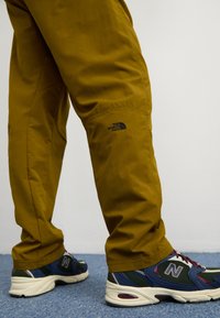 Khaki outdoorové kalhoty se syntetickou texturou, s logem The North Face na stehně, v kombinaci s modro-černými teniskami New Balance.