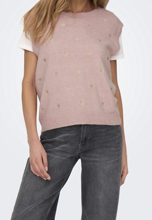 Woman wearing pink sleeveless sweater with small gold hearts over a white short-sleeve shirt and dark gray jeans standing with one hand down.