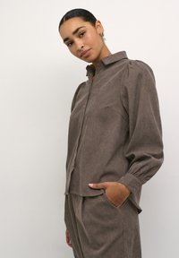 Brown button-up shirt with a collar, featuring puffed sleeves, textured fabric, and a relaxed fit. Pocket detail on the left side visible.
