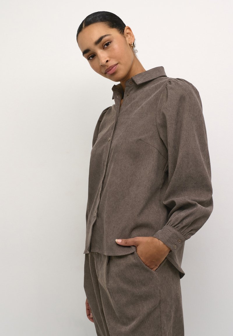 Brown button-up shirt with a collar, featuring puffed sleeves, textured fabric, and a relaxed fit. Pocket detail on the left side visible.
