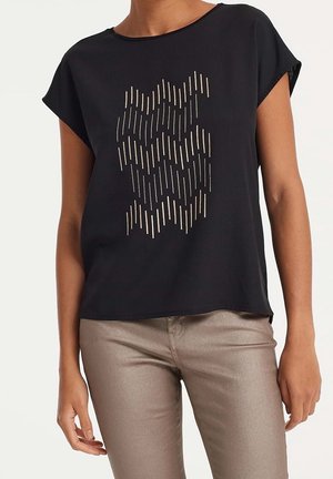 Kobieta ubrana w czarną koszulkę z krótkim rękawem z pionowym złotym wzorem kreskowym oraz metaliczne jasnobrązowe spodnie, stojąca na jednolitym tle.