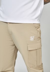 Beiga cargobyxor med sidofickor, med en vit broderad logo och ett mjukt, lätt tyg. Bärs med en vit t-shirt.