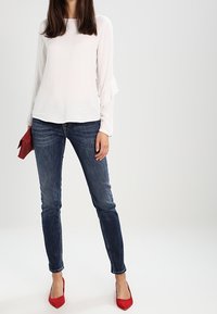 Vit långärmad blus med volanger vid ärmsluten, bärs med mörkblå skinny jeans och ljusröda pumps med spetsig tå, håller en röd kuvertväska.
