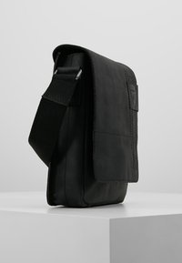 Borsa a tracolla in pelle nera con forma rettangolare, tracolla regolabile, pattina frontale, cuciture sottili e una piccola etichetta con logo.