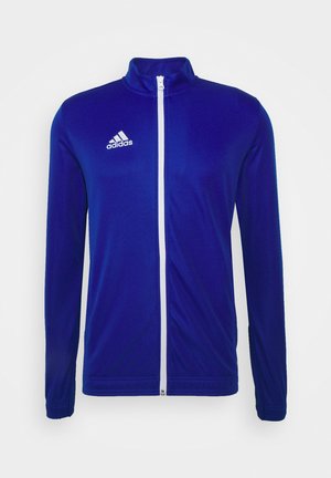 Plava sportska jakna izrađena od glatkog materijala. Ima visoki ovratnik i zatvarač punog dužine, s bijelim detaljima i Adidas logotipom na prsima.