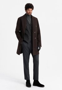 Dark brown overcoat, gray textured vest, gray turtleneck, black trousers, and black suede ankle boots. Gloves on one hand, neutral background.