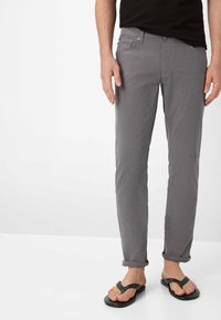 Mann trägt hochgekrempelte graue Hose und schwarze Flip-Flops, steht vor einem schlichten weißen Hintergrund.