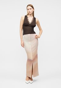 Ženska, oblečena v črno brezrokavnik z čipkasto izrezom, dolgo bež gradientno gumbasto krilo in bele koničaste čevlje, stoji z eno roko na boku.