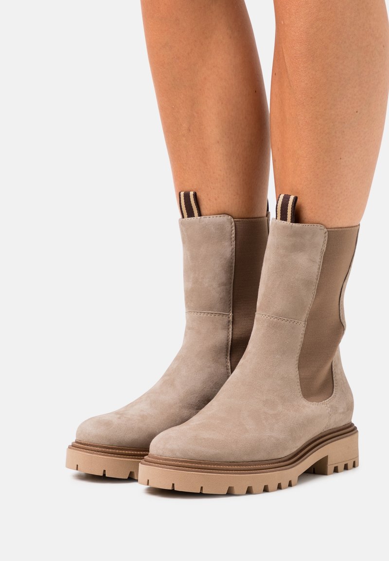 Bottes beige en daim mi-mollet avec des semelles en caoutchouc épaisses, des languettes rayées et des panneaux latéraux élastiques portées sur jambes nues.