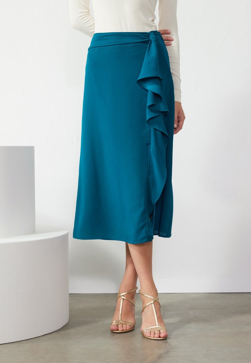 Femme portant une jupe midi turquoise avec des détails à volants et des sandales à talons hauts dorées à brides, debout sur un sol gris.