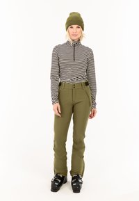 Gestreepte zwart-witte top met lange mouwen, groene broek met knoopdetails en een groene gebreide muts; staand in skibotten, neutrale achtergrond.