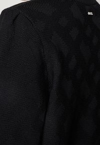 Suéter de punto negro con un patrón texturizado. Entre los detalles destacados se incluyen un cuello acanalado y un pequeño logo cerca del cuello en la parte posterior.